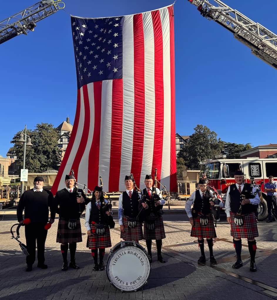 Virginia Highlands Pipes and Drums standing under huge US flag suspended between two firetruck ladders